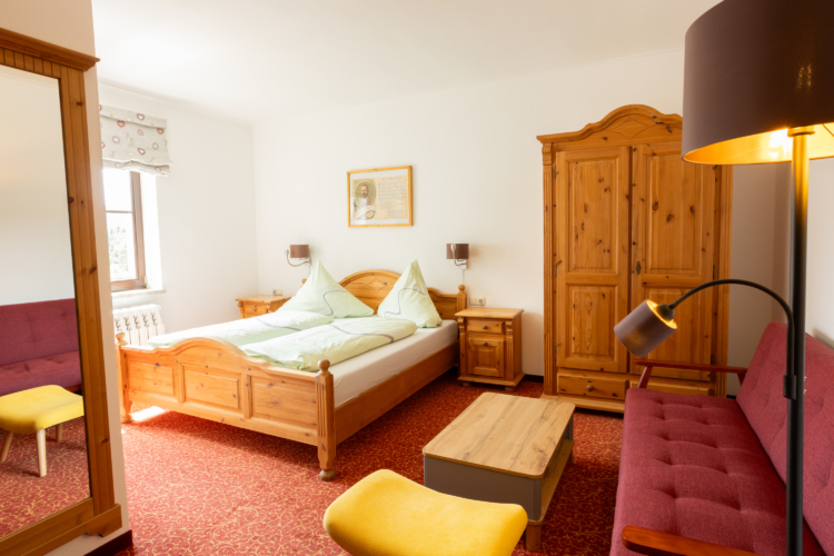 Berggasthof und Hotel Neues Haus - Doppelkomfortzimmer Ein Zimmer mit einem Doppelbett und einem Kleiderschrank. Dazu zwei kleine Nachtschränke. Die Möbel sind aus Holz. Es ist ein rotes Sofa, ein gelber Hocker, ein Holz-Tisch und eine Stehlampe zu sehen.